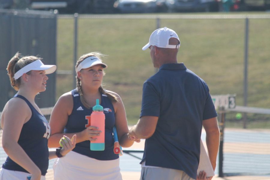 A male coah in a blue shirt and white hat talks to two female students, wearing white visors, blue shirts, and white skirts, during a tennis match.