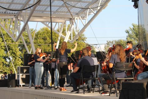 A woman in a blue shirt and black pants conducts a group of high school students with orchestra instruments.