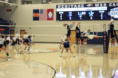 Two HHS volleyball players jump near the net with their hands raised. Behind them you can see the scoreboard where Southside leads 2-1. 